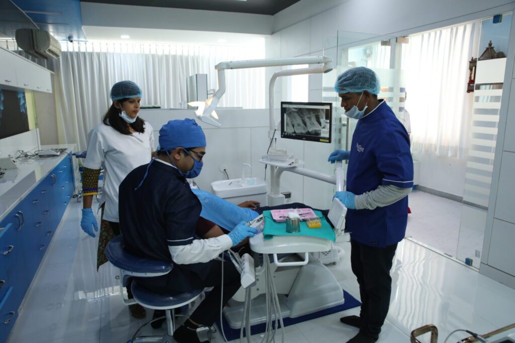Dental team treating a patient in a bright clinic; dentist at the chair with assistants nearby and a monitor showing an X-ray on the wall.