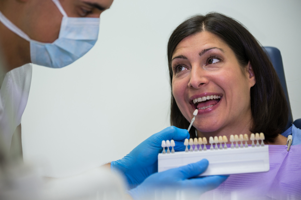 Dentist in a mask and blue gloves compares a tooth shade guide to the patient's smile in a dental chair.